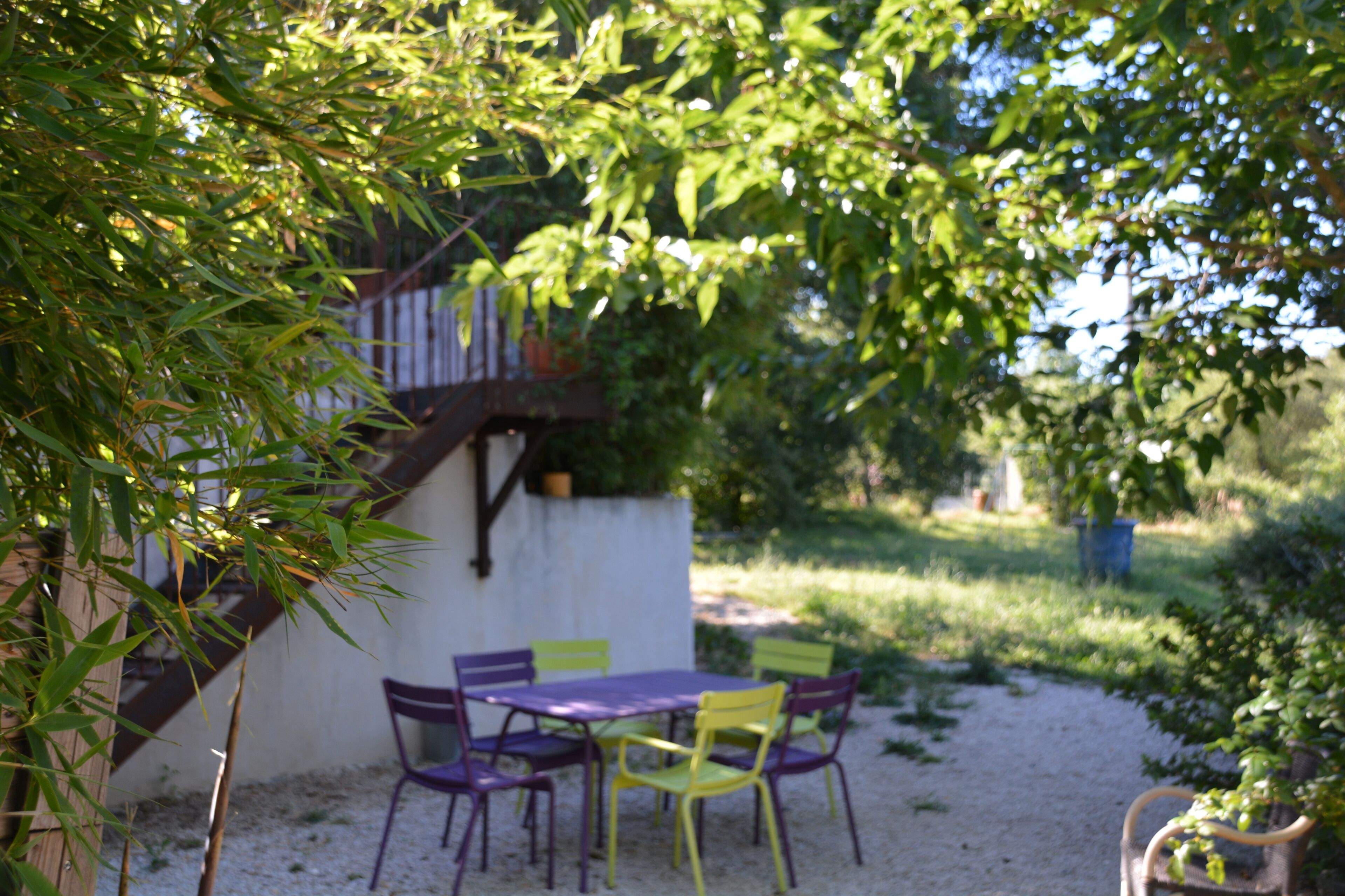 An outdoor patio with a table and chairs, surrounded by greenery and a white wall.