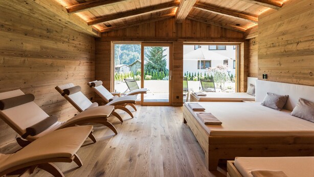 Interior de una cabaña de madera con una cama, sillones de descanso y un ventanal grande.