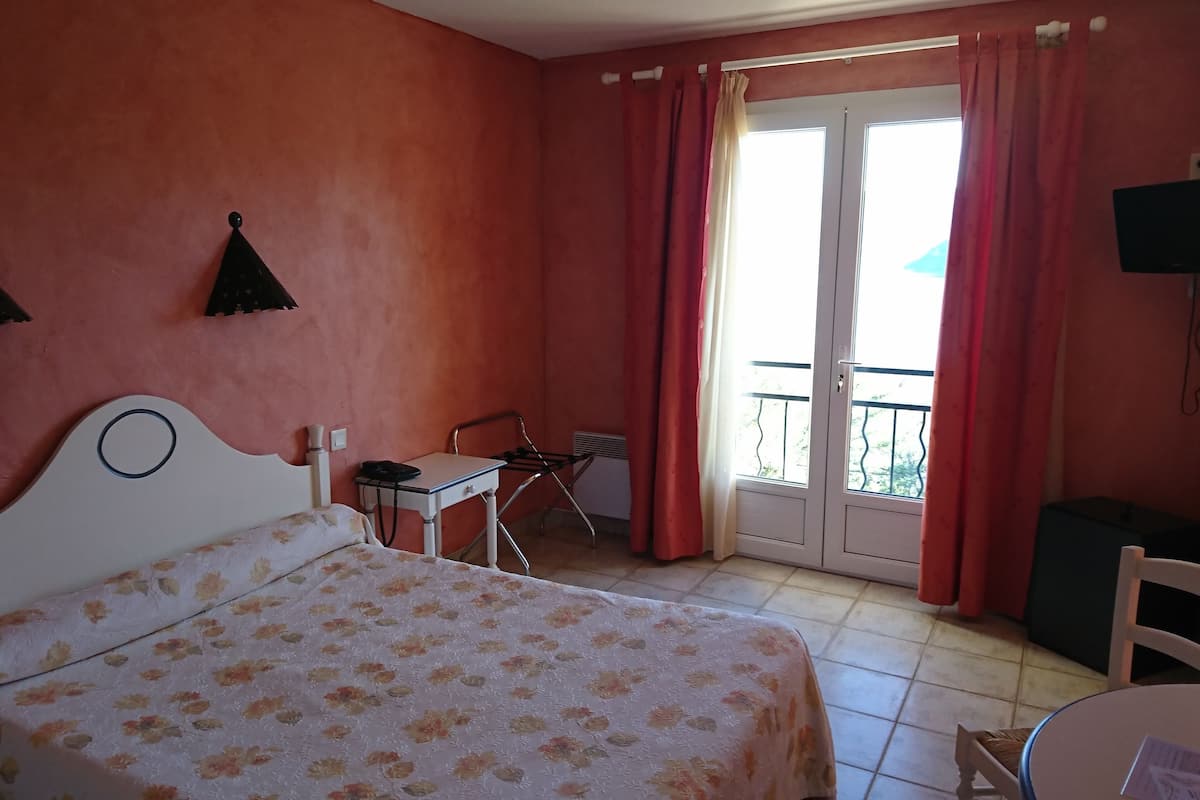 Un dormitorio con cama, escritorio, silla, calefactor y balcón con cortinas rojas.