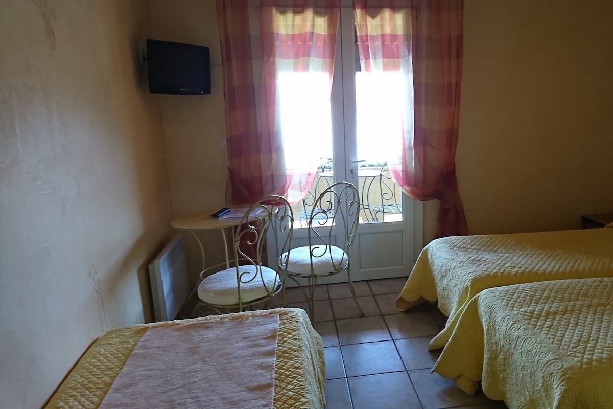 Una habitación con dos camas, una mesa pequeña con una silla, un televisor montado en la pared y una ventana con cortinas.