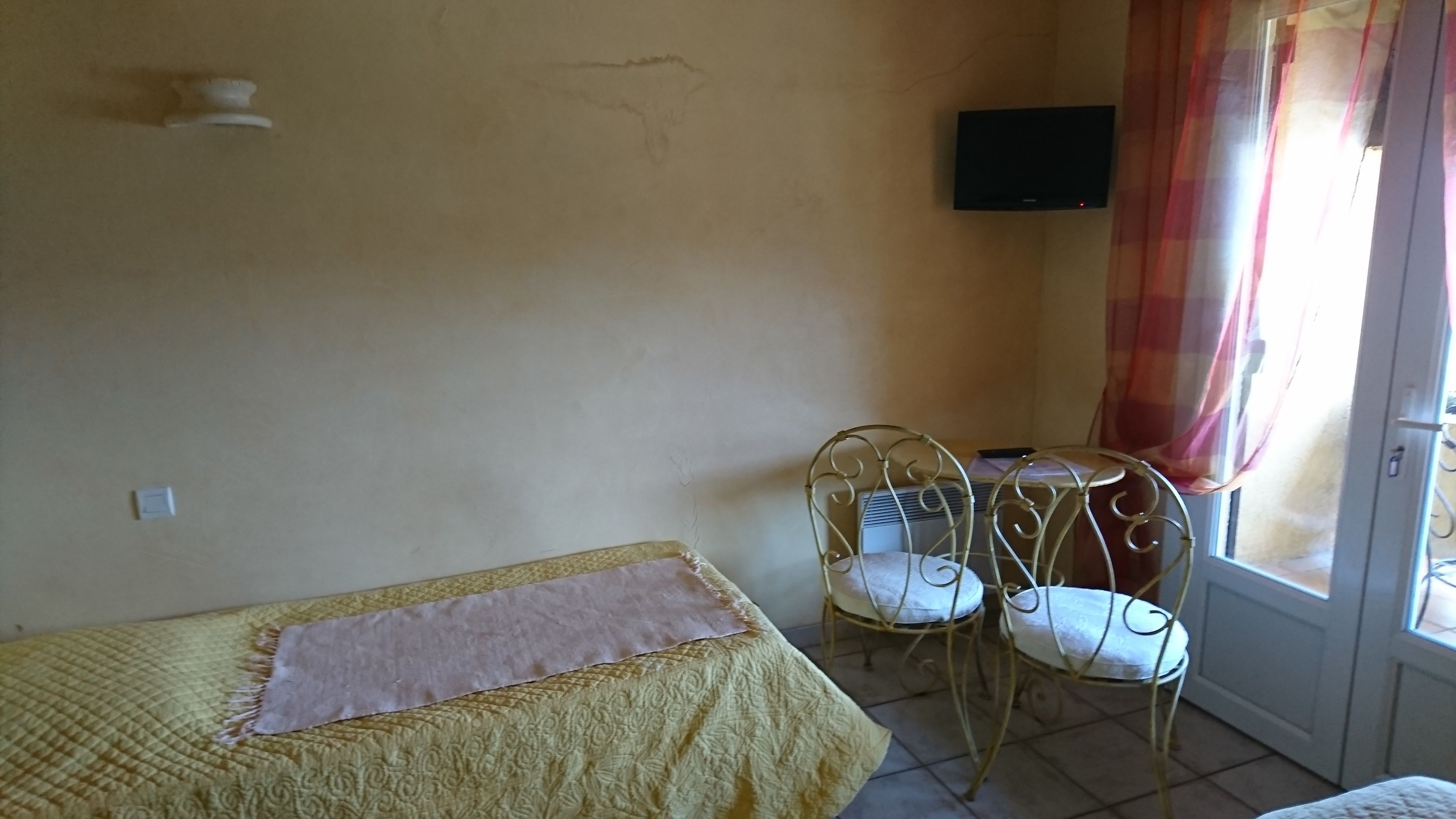 Habitació petita amb un llit, una taula, dues cadires, una televisió i una finestra amb cortines.