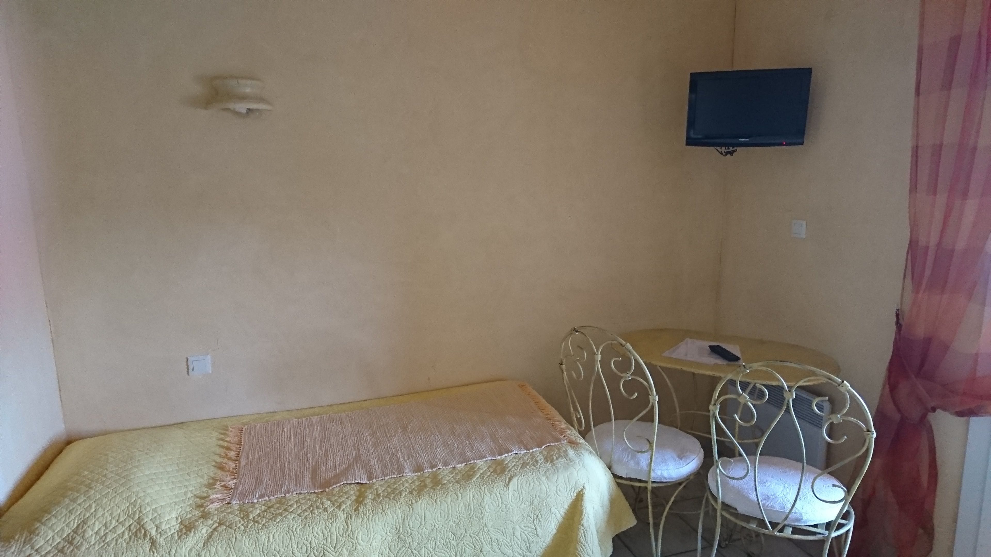 Una habitació petita amb un llit, una taula i dues cadires, un televisor fixat a la paret i una cortina.