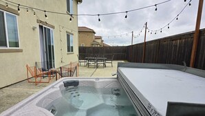 Un jacuzzi en un patio trasero con luces colgantes, una valla de madera y mobiliario exterior.