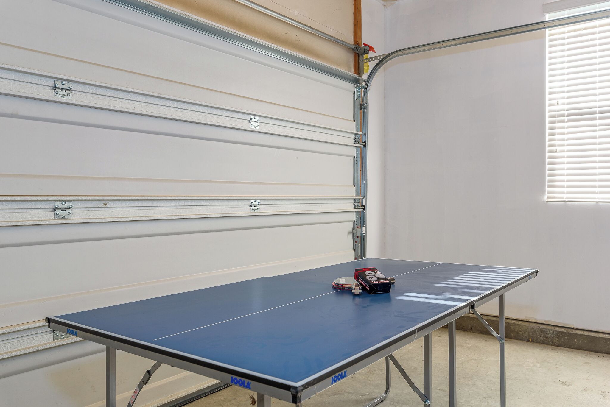 Une table de ping-pong dans une pièce dotée d’une porte de garage et d’une fenêtre avec des stores.