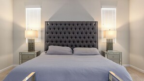 Un dormitorio con un cabecero capitonado, dos mesitas de noche con lámparas, una cama con colcha azul y una ventana con persianas.