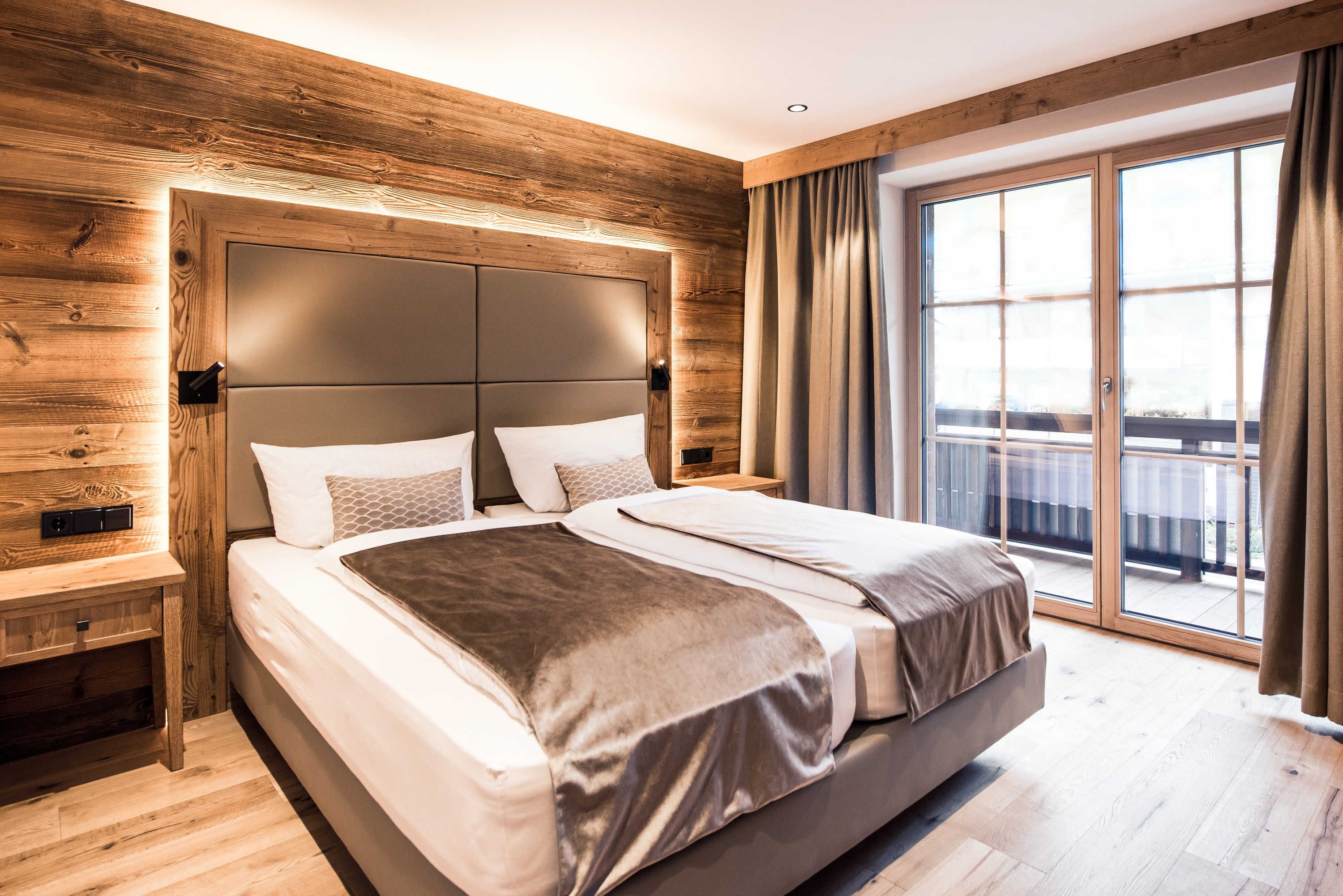 A modern bedroom with a large bed, wooden paneling, a sliding glass door to a balcony, and a nightstand.