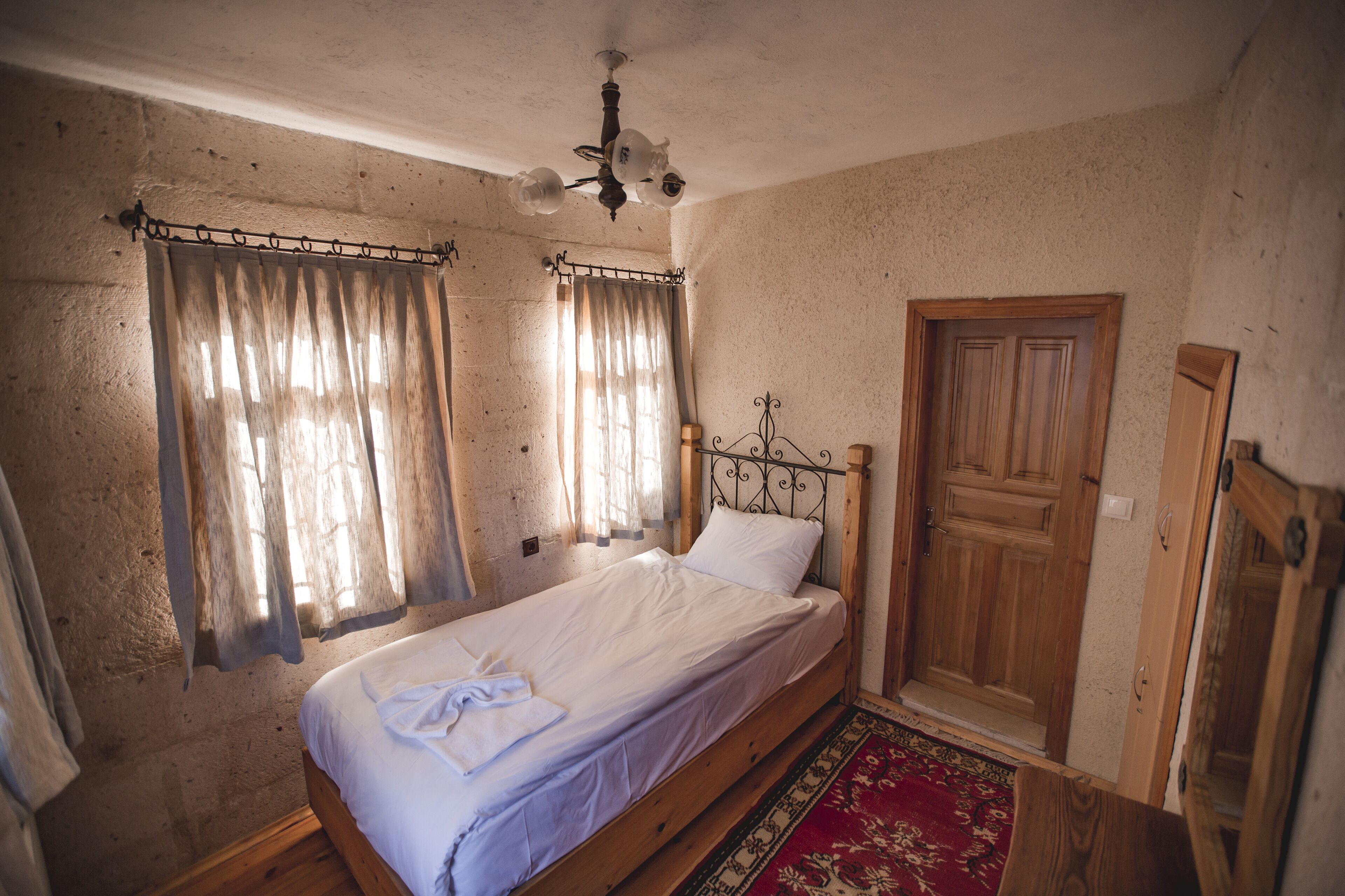 Una cama individual con cabecera de madera, una alfombra roja estampada y una puerta de madera.