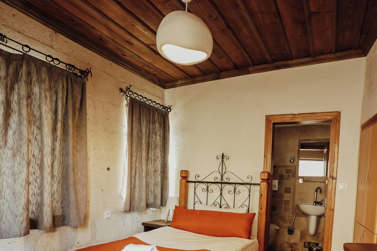 A wooden ceiling, a bed with an orange bedspread, a patterned rug, and a bathroom with a sink and mirror.