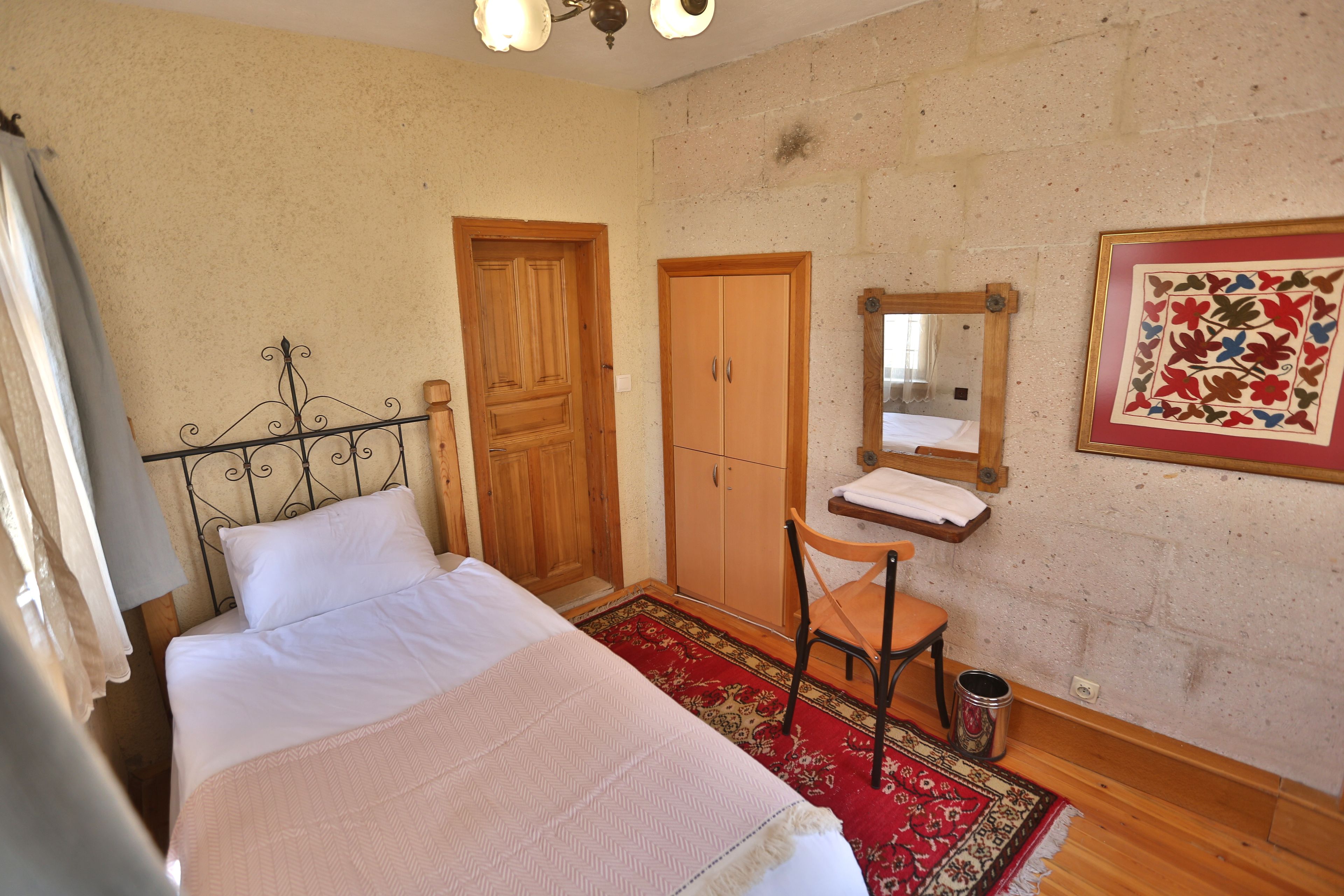 A bedroom with a bed, wooden wardrobe, a small wooden chair, a red patterned rug, and a wall-mounted mirror.