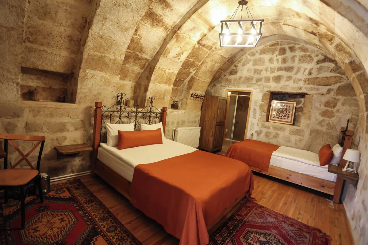 A rustic room with a vaulted stone ceiling, a wooden bed with orange bedding, a wooden chair, and a small table.