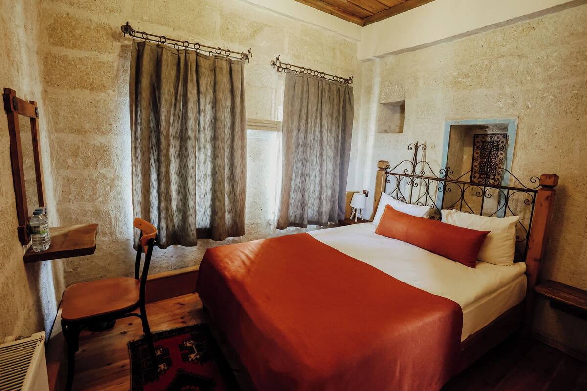 A bedroom with a wooden bed, a chair, a shelf with a bottle, and a window with curtains.