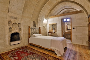 Quarto com lareira de pedra, cama com colcha branca, piso de madeira e tapete vermelho estampado.