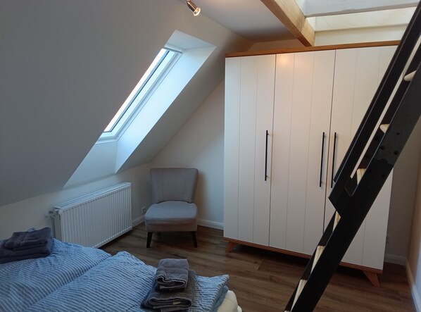 Un dormitorio con techo inclinado, un tragaluz, una cama con ropa de cama azul, una silla y un armario.