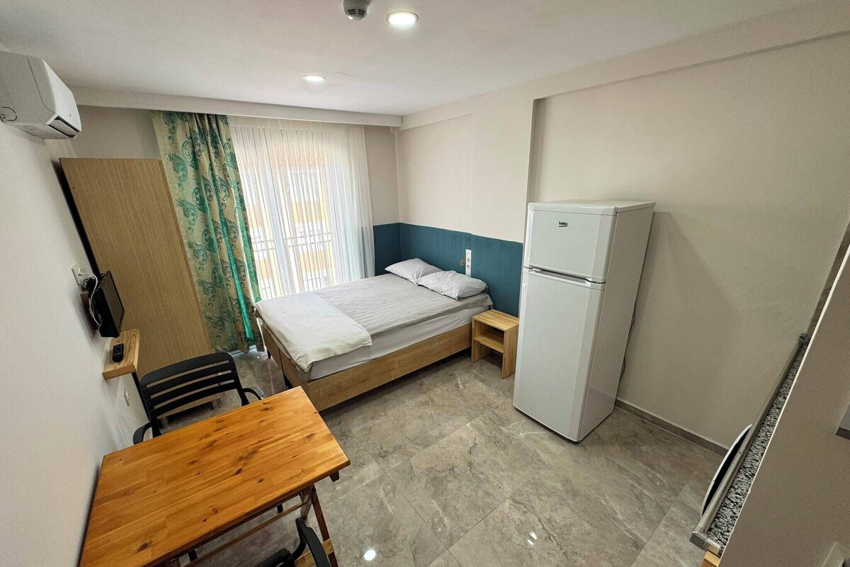 Una habitación pequeña y limpia con cama, escritorio, silla, refrigerador y ventana con cortinas.