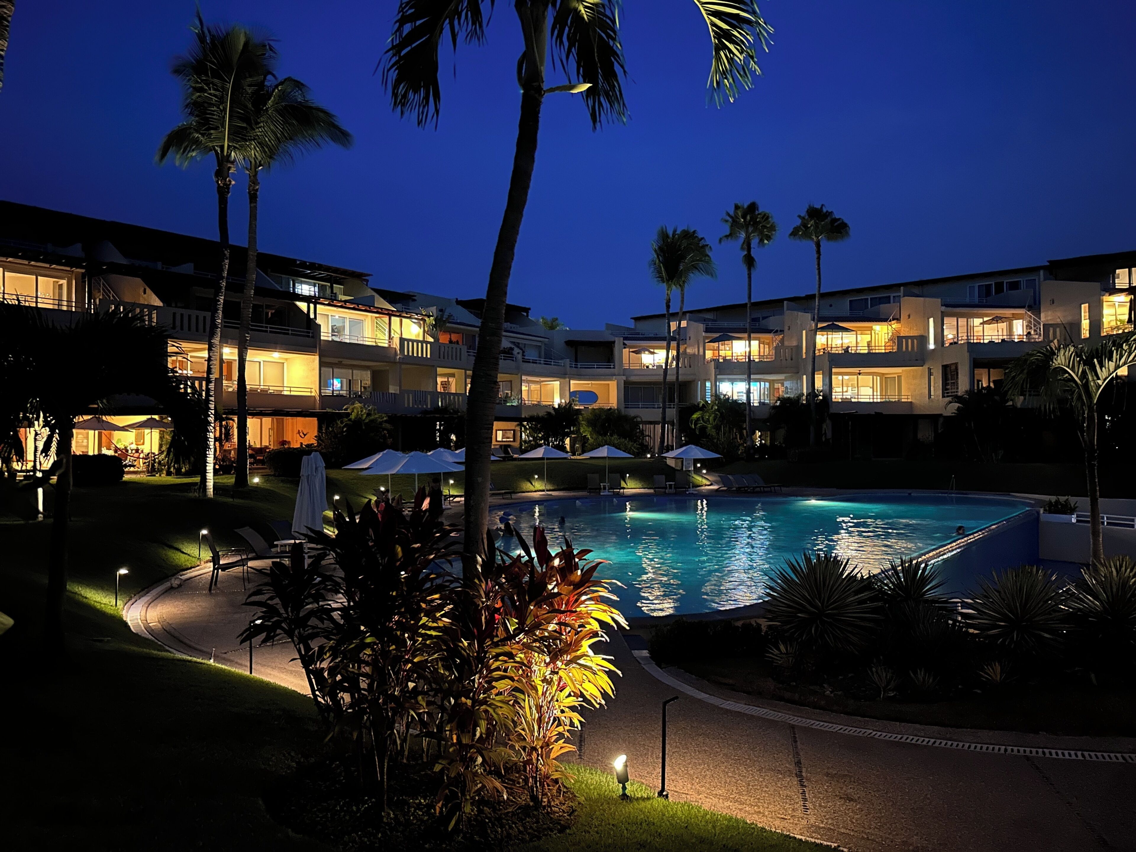 A resort with a swimming pool, palm trees, and illuminated buildings at night.