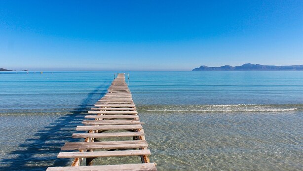 Eine hölzerne Pier, die sich in ruhiges blaues Wasser erstreckt und zu einer entfernten Küstenlinie mit sanften Hügeln führt.