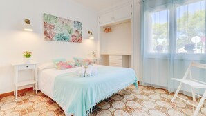 Ein Schlafzimmer mit einem Bett, einem Nachttisch, einem Sessel, einem Fenster mit Vorhängen und einer Wand mit einem Blumenbild.