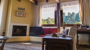 A cozy living room with a fireplace, a television, and a view of rocky mountains through large windows. - Tasos House_Panoramic View  (Kalabaka)