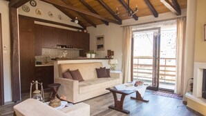 A spacious living room with a sofa, coffee table, and a fireplace. There is a kitchen area with wooden cabinets and a tiled backsplash. - Tasos House_Panoramic View  (Kalabaka)