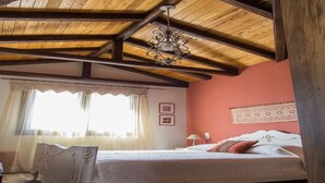 A bedroom with a wooden ceiling, a bed with white bedding, a nightstand, and a window with curtains. - Tasos House_Panoramic View  (Kalabaka)