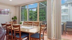 Et spiseområde med et træbord og stole, store vinduer med udsigt til grønne områder, og en balkon med udsigt til en bygning.