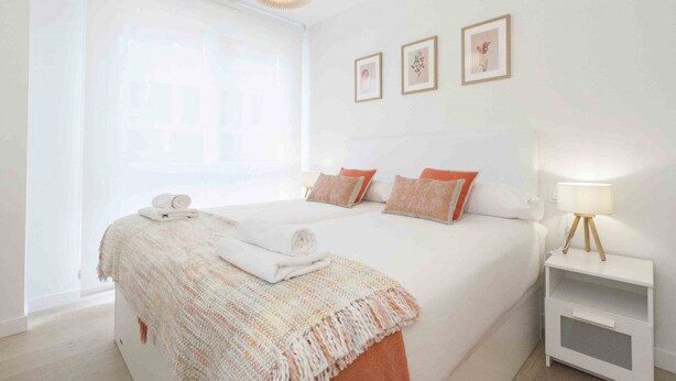 A hotel room with a large bed, a bedside table with a lamp, and a window with blinds. - Seattle | The Rentals Collection (San Sebastián)