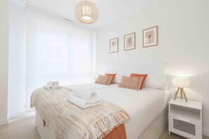 A hotel room with a large bed, a bedside table with a lamp, and a window with blinds. - Seattle | The Rentals Collection (San Sebastián)