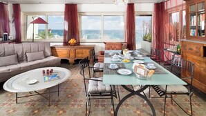 A spacious living room with a large window overlooking the sea, a round coffee table, a sofa, and a dining area with a table set for a meal.