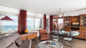 A living room with a sofa, a round coffee table, a dining table with chairs, a red chair, a wooden cabinet with books, and a view of the sea through large windows.