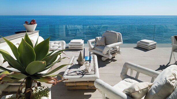 A deck with white outdoor furniture, a potted plant, and a view of the ocean.