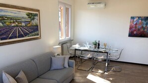 Ein Wohnzimmer mit einer Couch, einem Esstisch mit Stühlen, einem Fenster und einem Bild an der Wand.