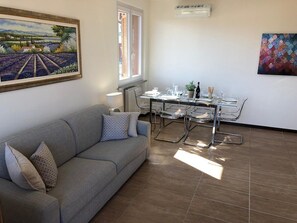 Ein Wohnzimmer mit einer Couch, einem Esstisch mit Stühlen, einem Fenster und einem Bild an der Wand.
