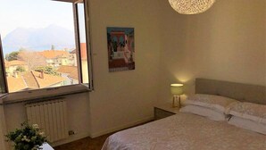 Ein Schlafzimmer mit einem Bett, einem Nachttisch, einer Lampe, einem Fenster mit Blick auf Gebäude und Berge sowie einem Poster an der Wand.