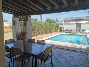 A covered outdoor area with a pool, lounge chairs, and a dining table with chairs.