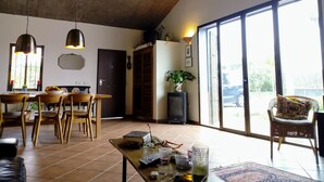 Amplia sala de estar con techo alto, mesa de comedor con sillas, una mesa de centro con diversos objetos y un gran ventanal corredera que da a un espacio exterior.
