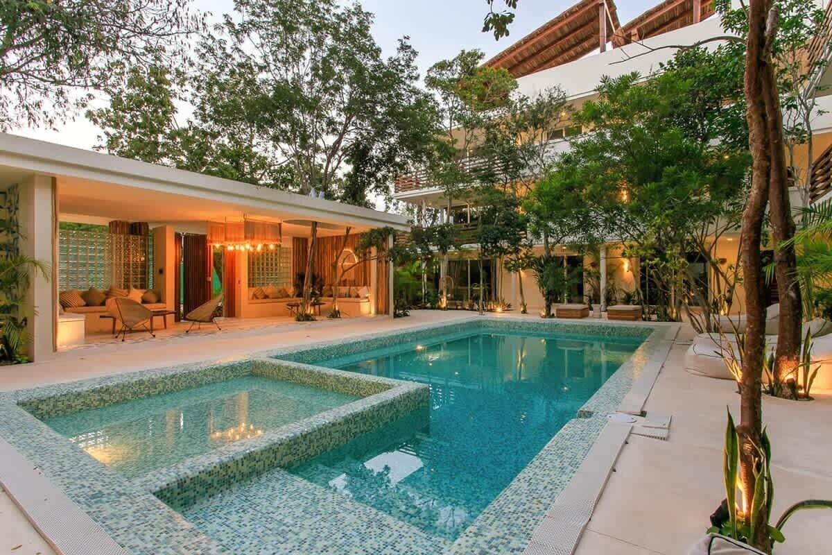 Une piscine dont le bord est revêtu de carrelage, entourée d’un espace de détente couvert, avec des chaises et un canapé.