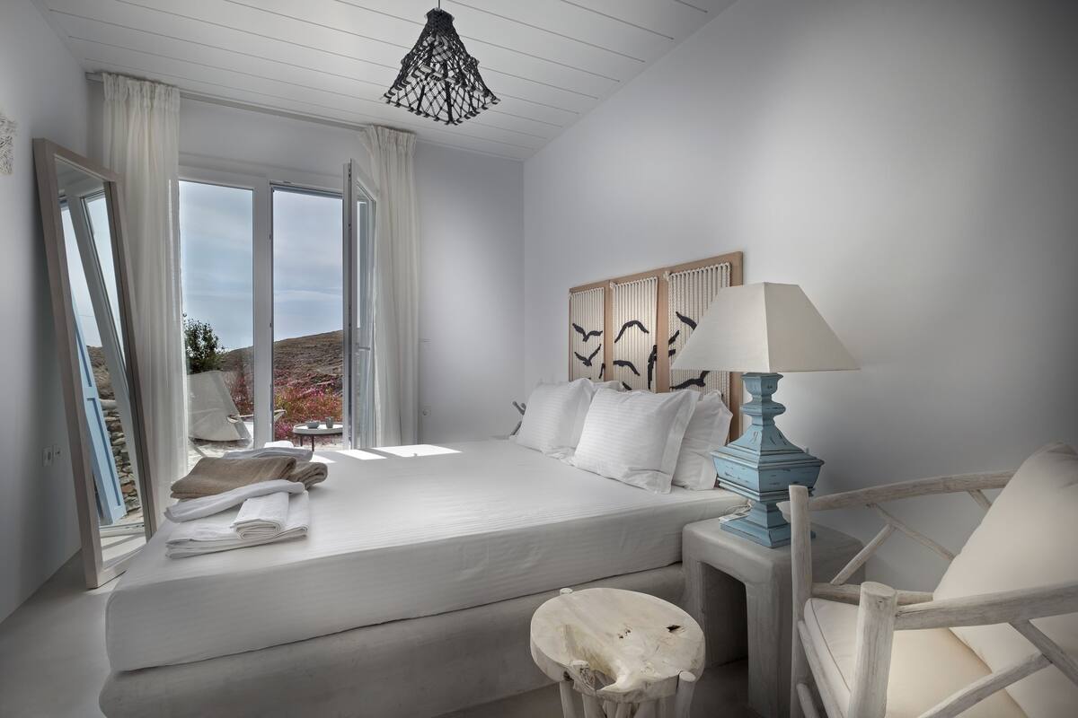 A bedroom with a large bed, a bedside table with a lamp, a chair, and a view of the outdoors.