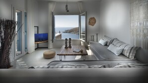 A modern living room with a large window offering a coastal view, a sofa with striped cushions, a wooden coffee table, and a flat-screen TV. - Simousi Villas (Kithnos)