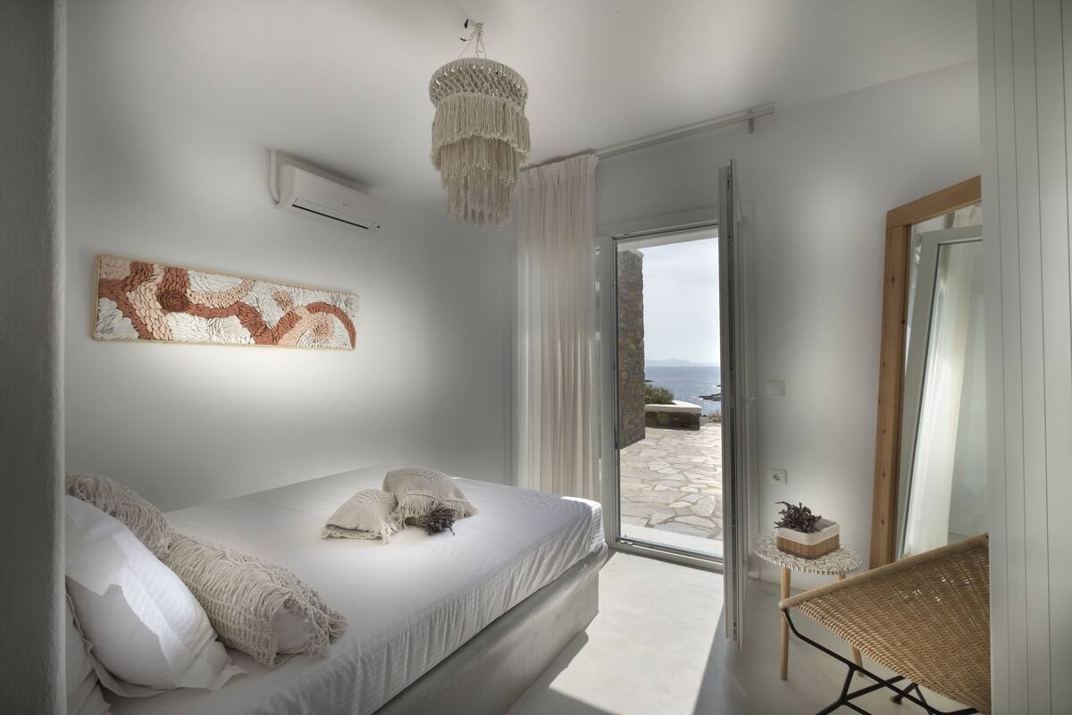 A bedroom with a bed, a chandelier, a view of the outdoors, and a wicker chair.