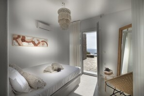 A bedroom with a bed, a chandelier, a view of the outdoors, and a wicker chair. - Simousi Villas (Kithnos)