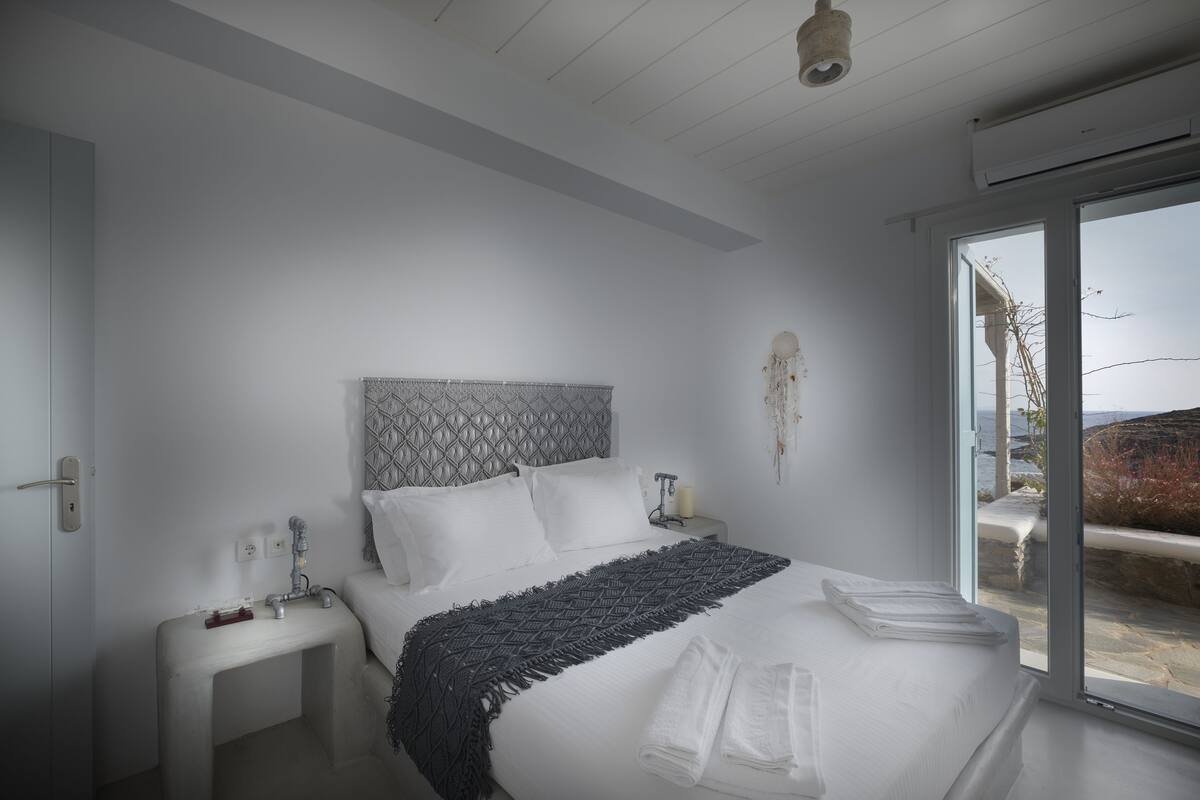 A modern bedroom with a large bed, a bedside table, a nightstand, and a view of the outdoors through a glass door.
