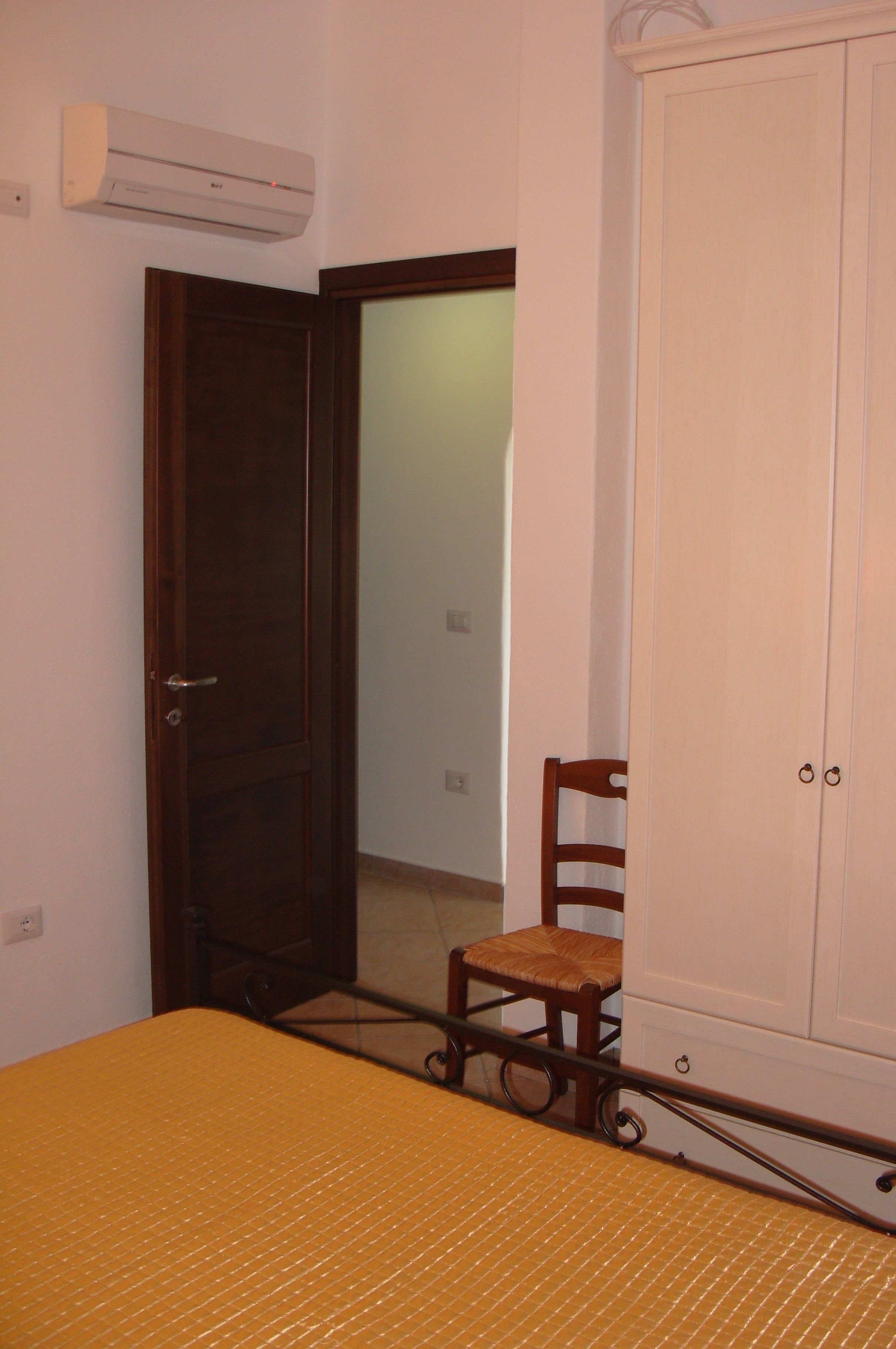 A room with a bed, a wooden chair, a wardrobe, and an air conditioning unit.