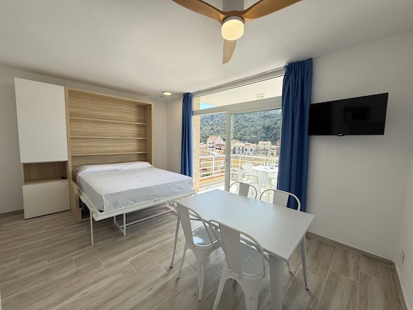 A bedroom with a bed, a table, chairs, a ceiling fan, a TV, and a window with curtains.