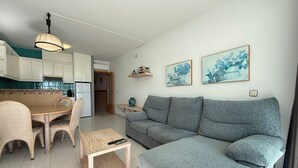 Ein modernes Wohnzimmer mit grauem Sofa, Korbstühlen, einem Holztisch und einer Küche im Hintergrund.