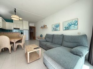 A modern living room with a grey sofa, wicker chairs, a wooden coffee table, and a kitchen in the background.