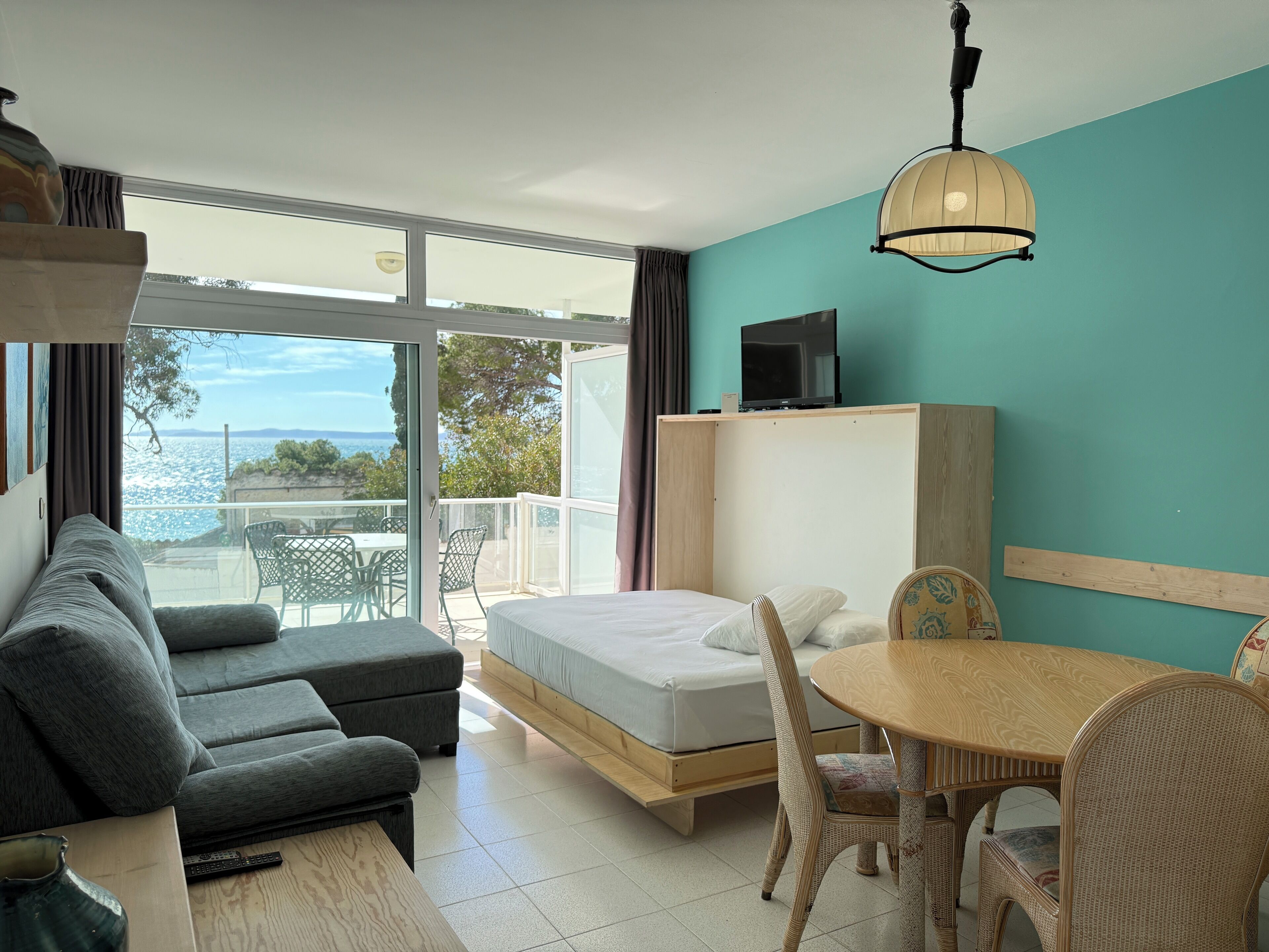 A modern hotel room with a balcony, a sofa, a bed, a dining table, and a television.