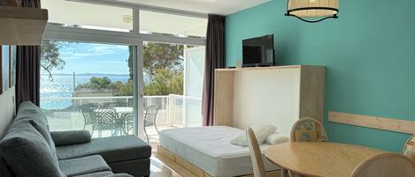 A modern hotel room with a balcony, a sofa, a bed, a dining table, and a television.