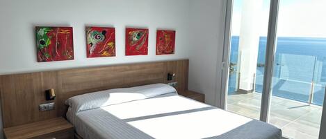Une chambre moderne avec un grand lit, une table de chevet, une commode et une vue sur l’océan grâce à une porte-fenêtre coulissante.