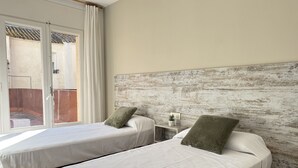 Quarto de hotel com duas camas de solteiro, cabeceira de madeira, mesa de cabeceira pequena com uma planta, e vista para um pátio por uma porta deslizante.