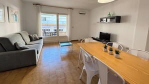 Um apartamento moderno com um sofá cinzento, uma mesa de jantar de madeira, cadeiras brancas, uma televisão e uma varanda com vista para edifícios.